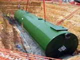 A large green underground storage tank being installed at a construction site with red safety barriers.