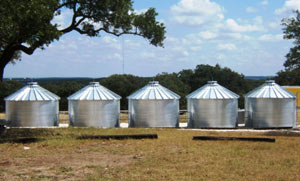 Five galvanized steel water storage tanks with conical roofs, aligned outdoors in a rural setting.