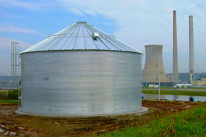 Large galvanized steel water storage tank with a conical roof, located in an industrial area with smokestacks in the background.